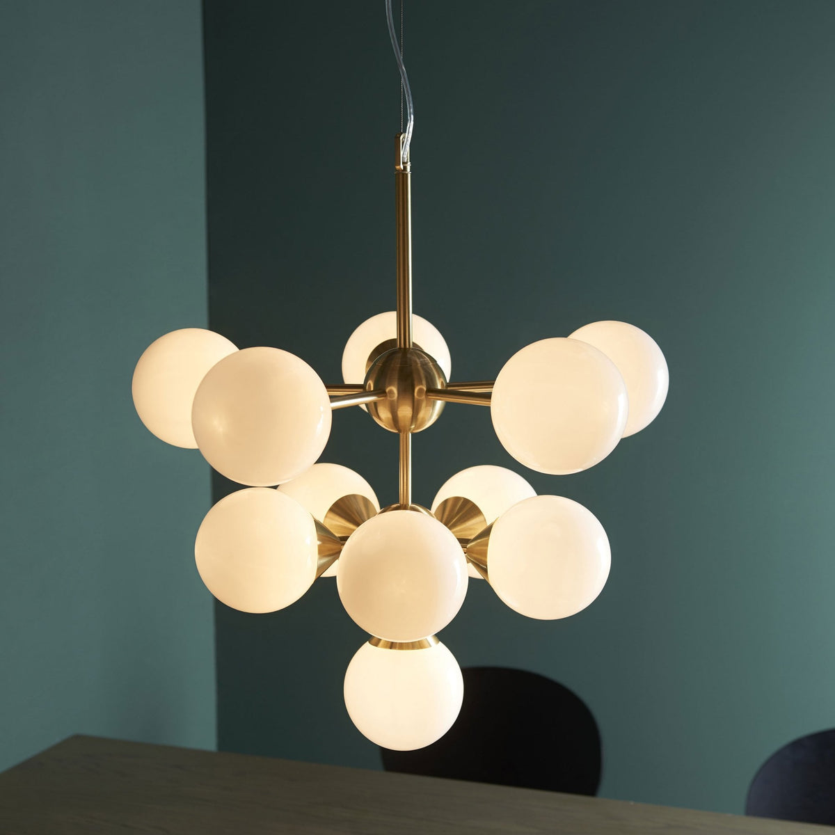 A brass chandelier with ten white globe lights hangs above a dining table in a room with teal walls.