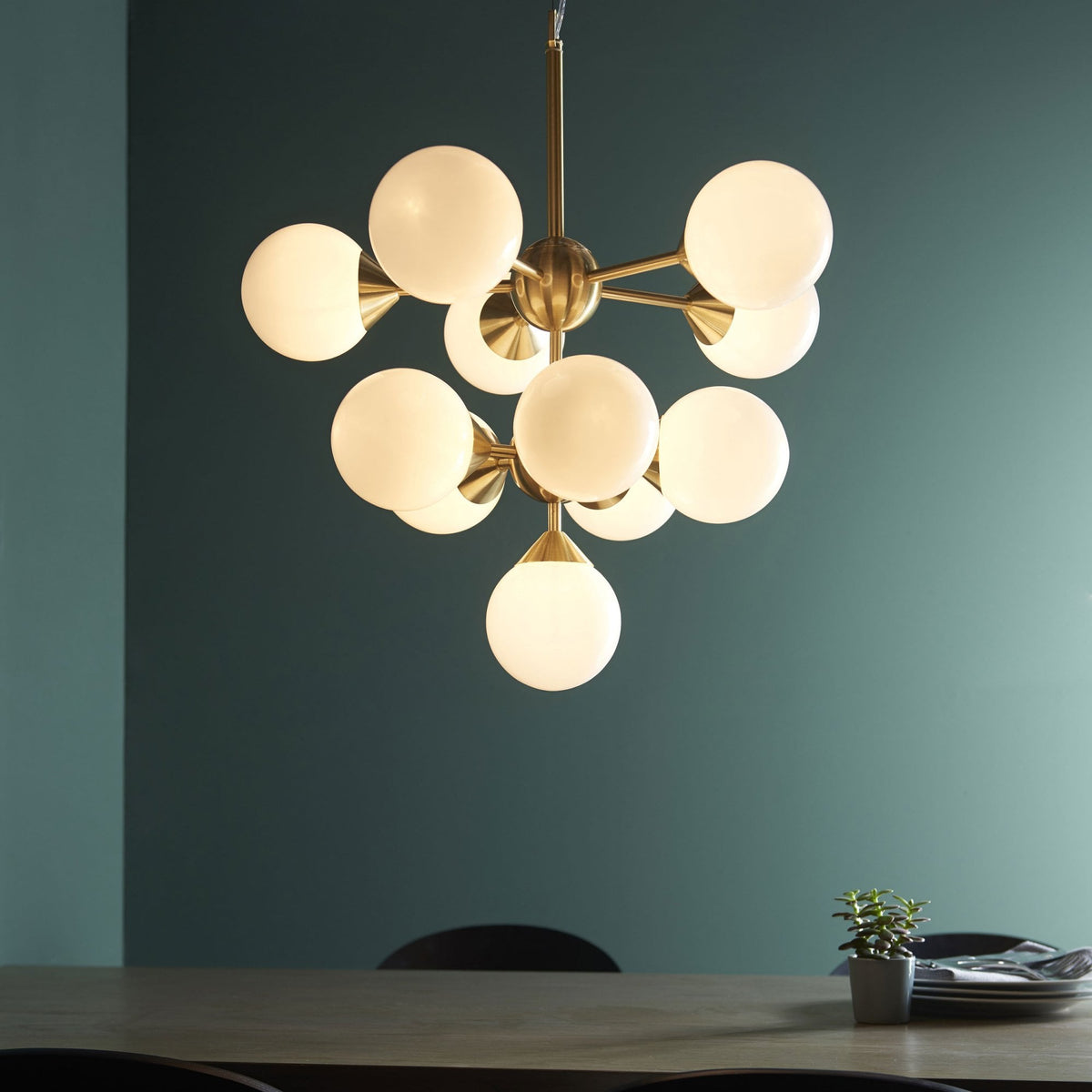 A brass chandelier with ten white globe lights hangs over a dining table with a small plant.