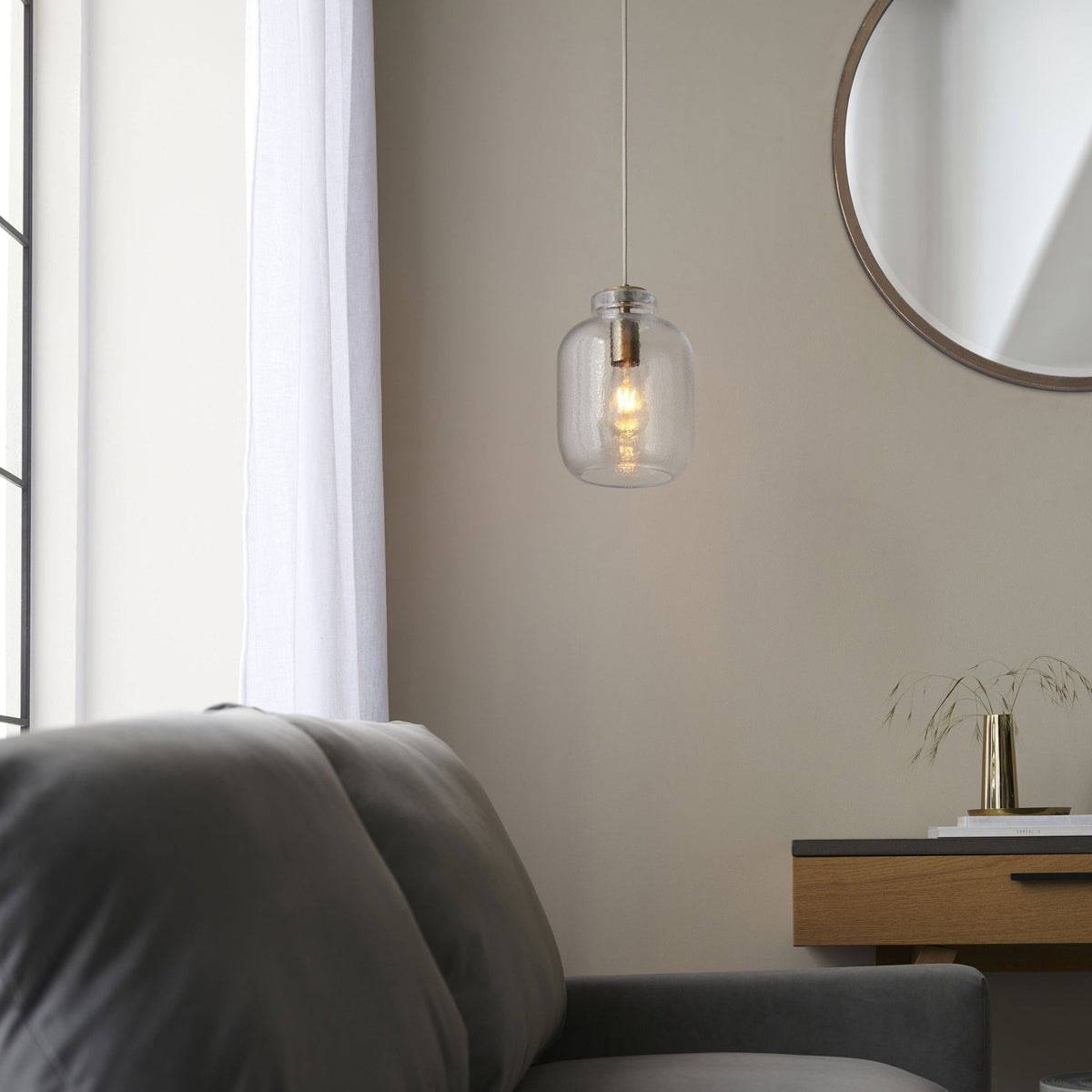 A hammered glass pendant light illuminates a modern living room with a grey sofa and mirror.