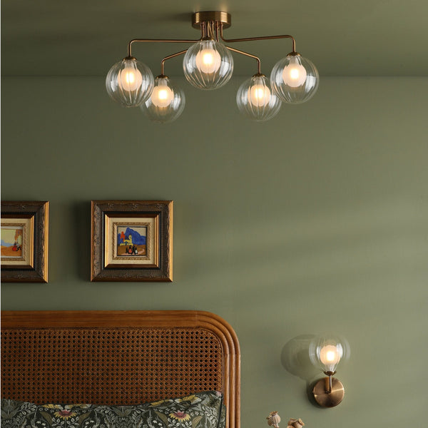 A bedroom with a green wall, a rattan headboard, and two framed pictures above it. A ceiling light with five glass globes and a wall sconce with a single glass globe illuminate the room.