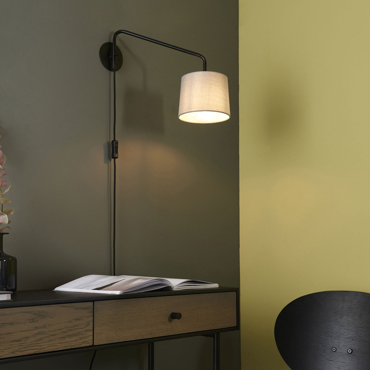Black wall light with a white fabric shade, illuminating a dark green wall above a wooden console table.