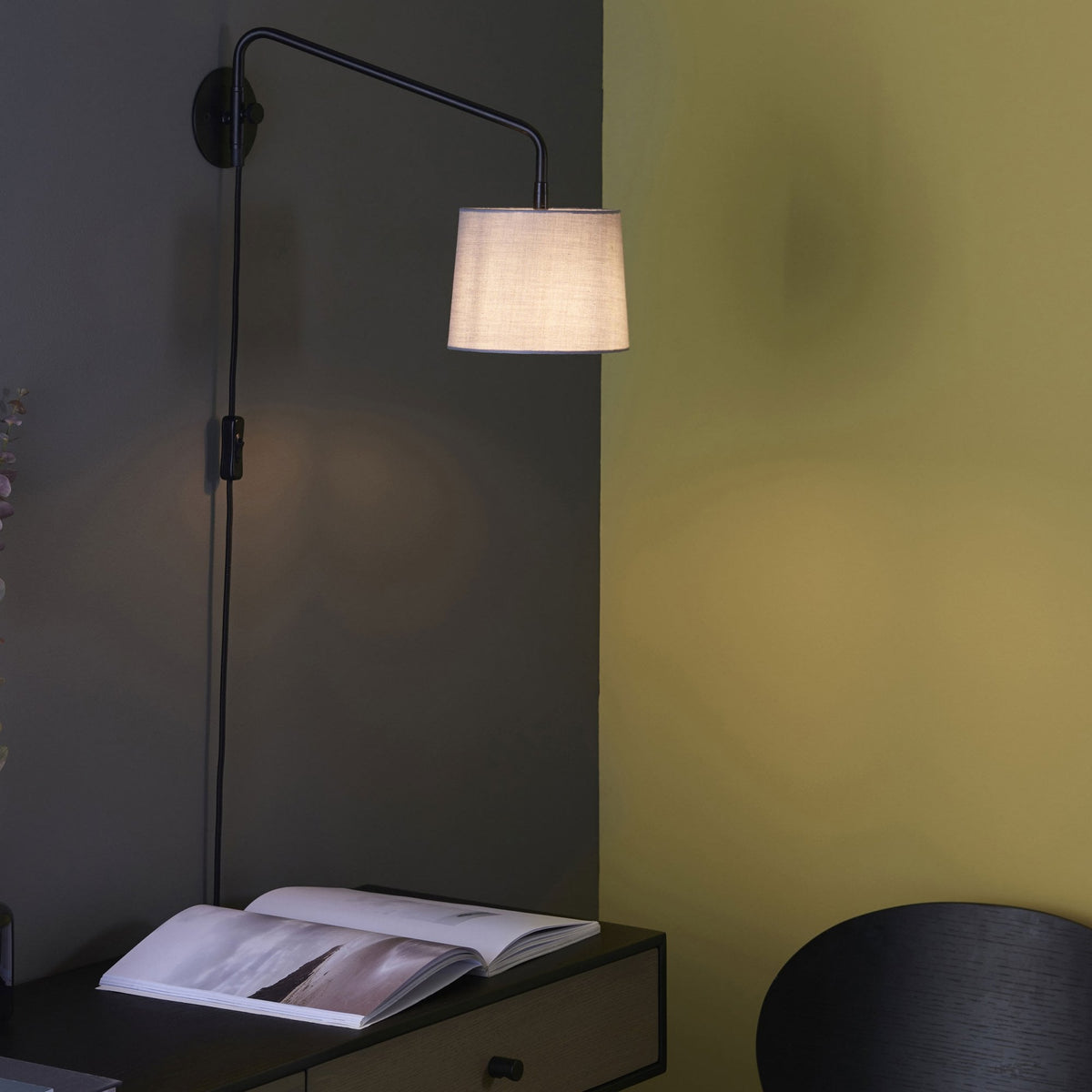 Black wall light with a white fabric shade illuminating a dark green wall above an open book on a table.