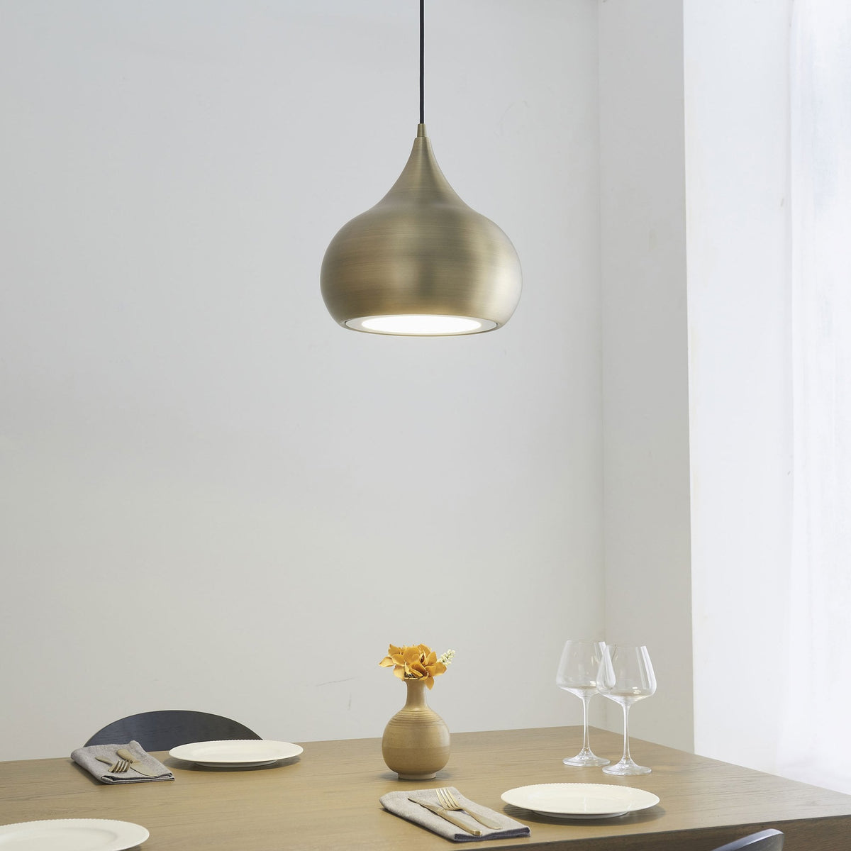 A brass pendant light hangs above a dining table set with plates, cutlery, and wine glasses.