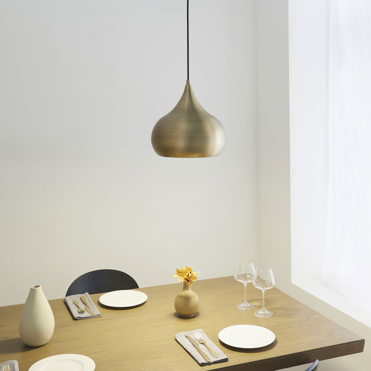 A brass pendant light illuminates a dining table with place settings and a small vase with flowers.