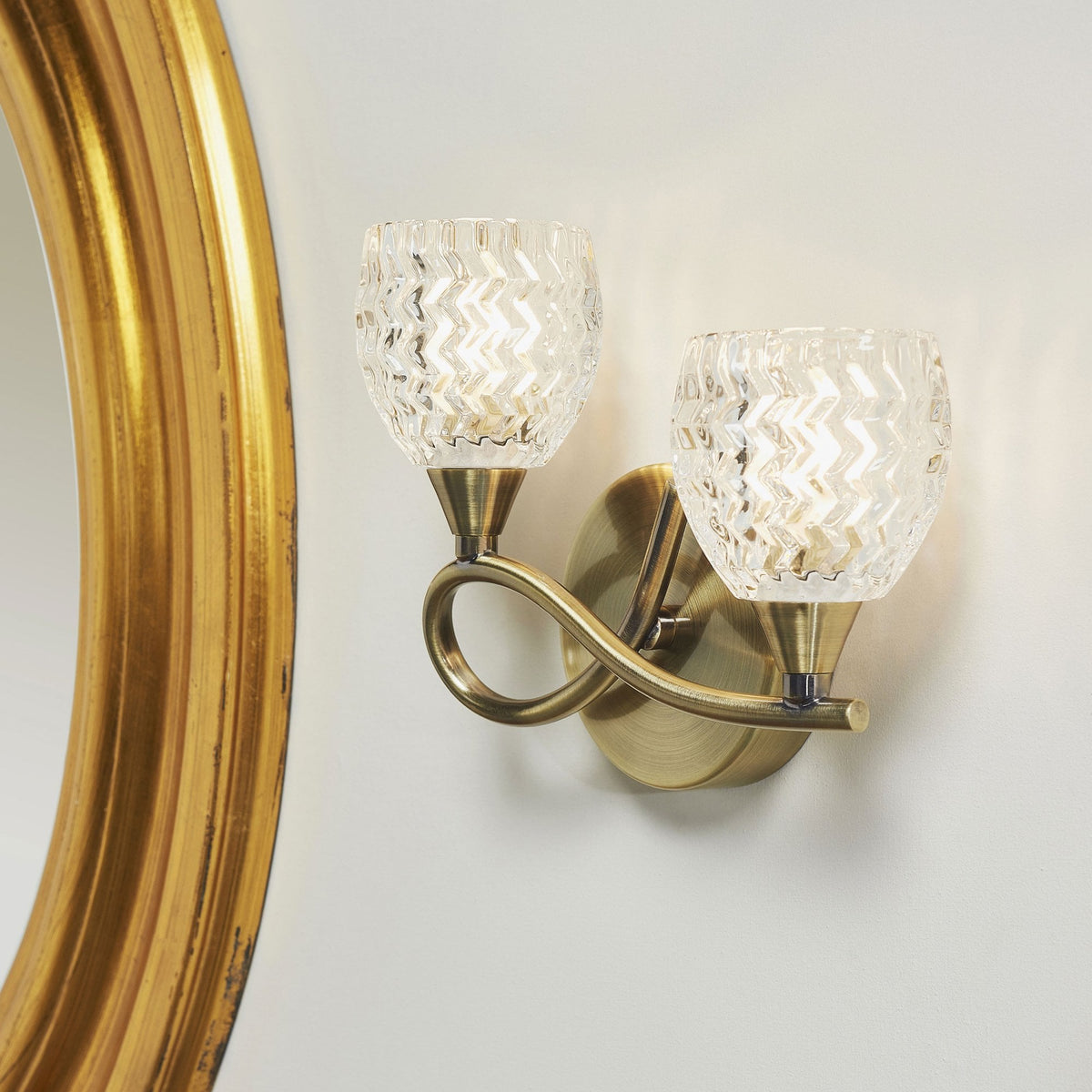 Two-light wall sconce with clear, textured glass shades and an antique brass finish next to a gold mirror.
