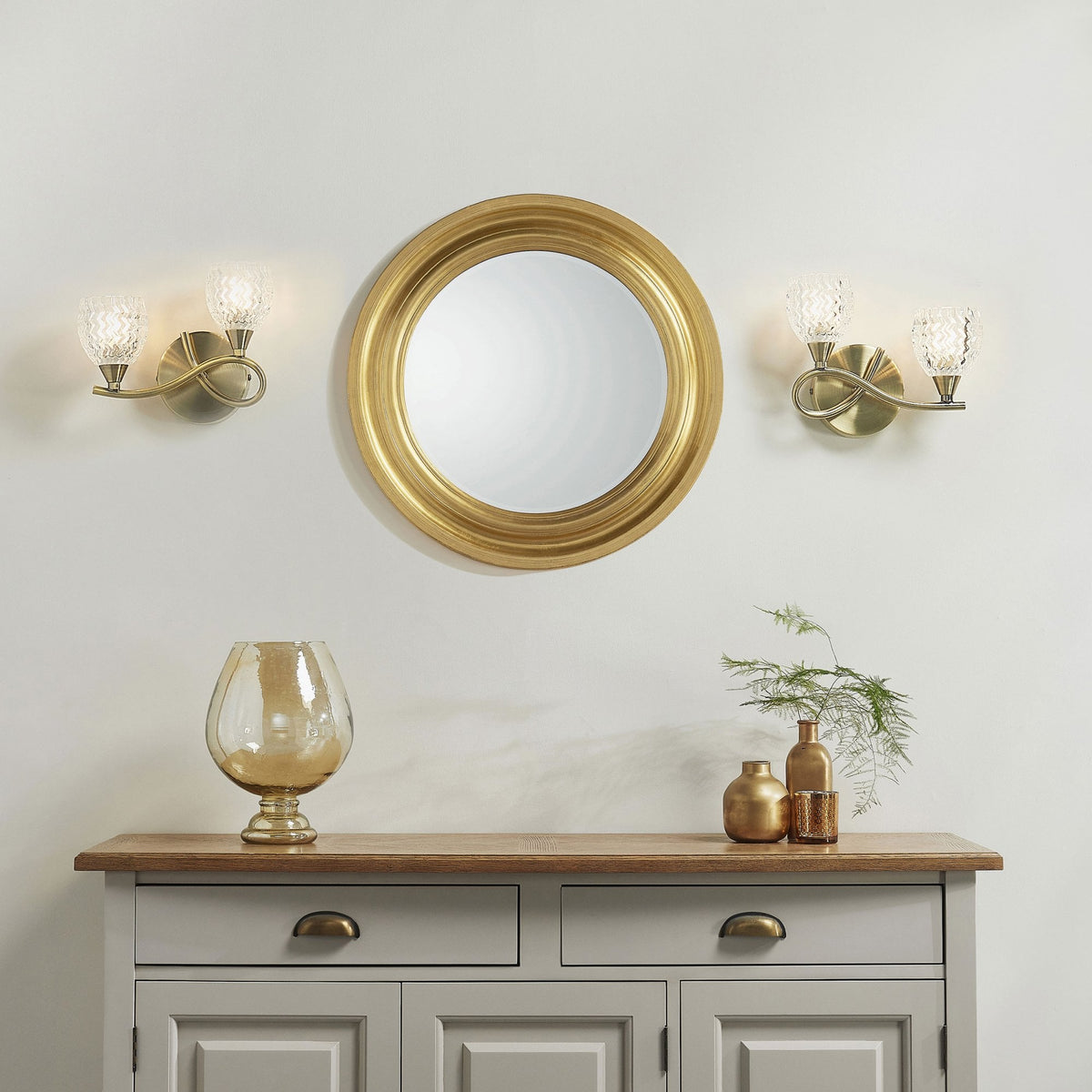 Two wall sconces flanking a round gold mirror above a gray console table with decorative items.