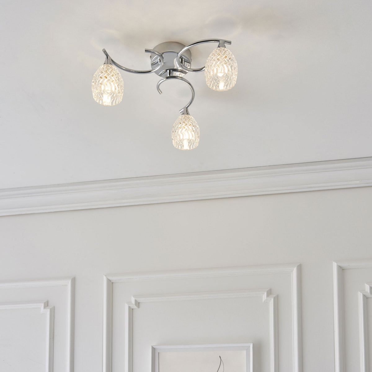 Three-light ceiling fixture with chrome arms and textured glass shades above a white paneled wall.