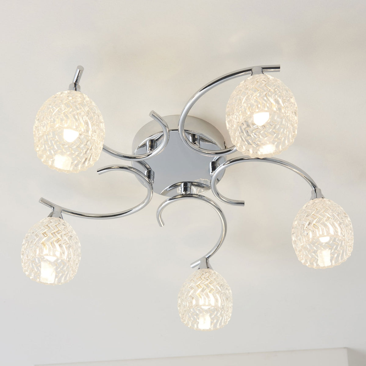 Close-up of a chrome ceiling light with five clear, textured glass shades, illuminated against a white ceiling.