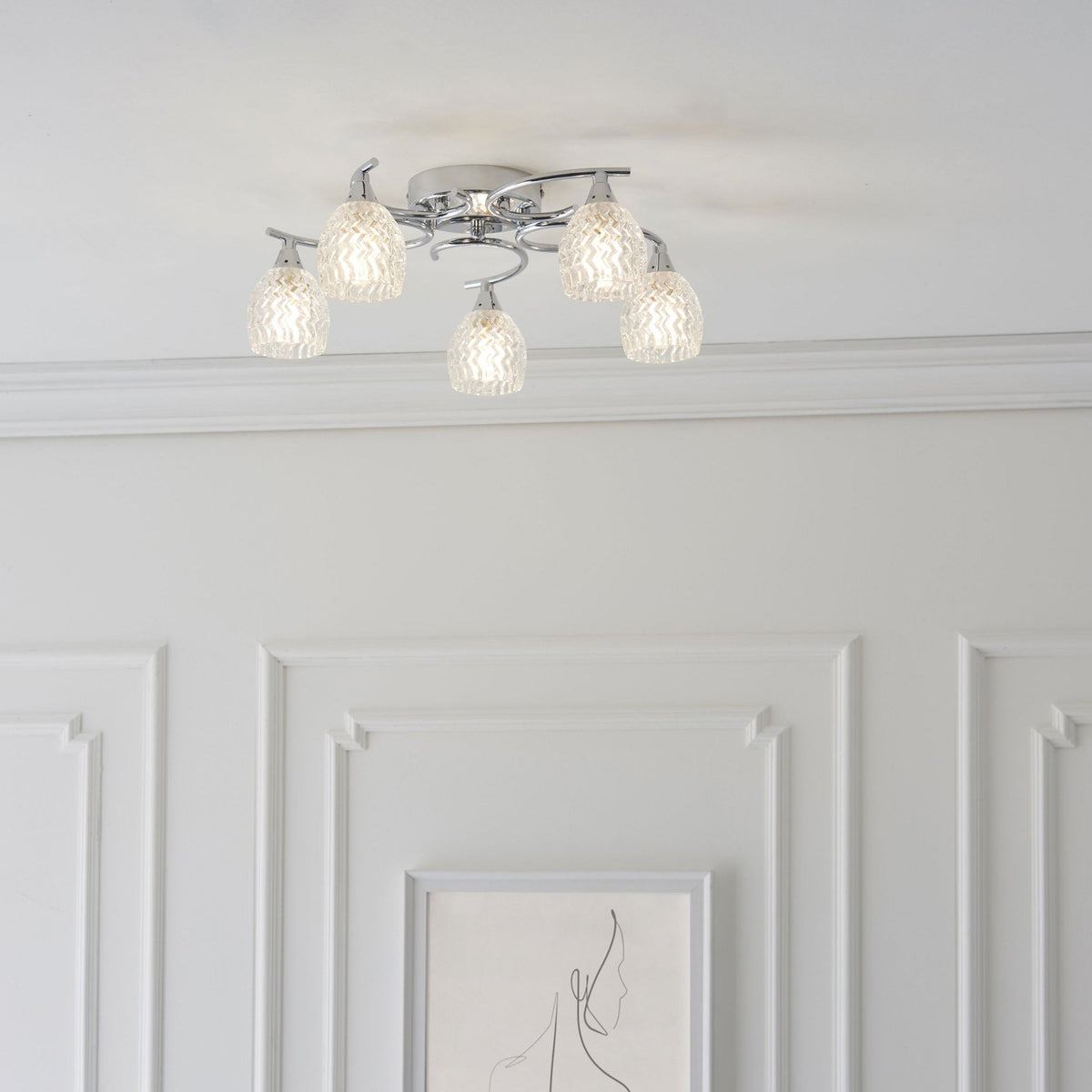 Chrome ceiling light with five clear, textured glass shades, illuminated above a white wall with paneling.
