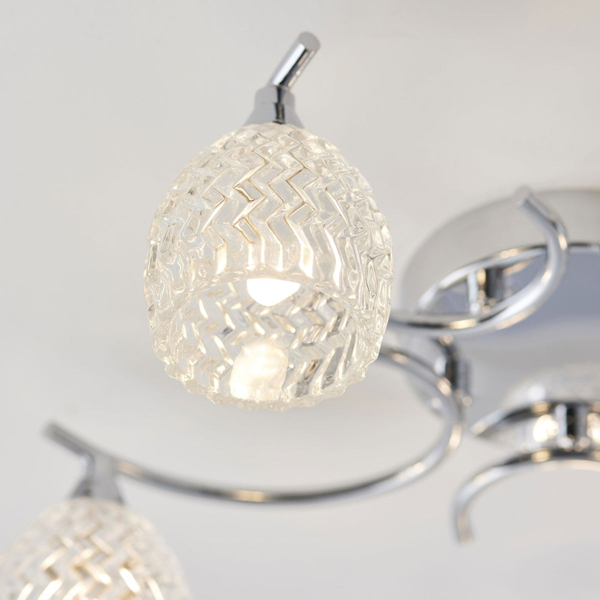 Close-up of an illuminated, clear, textured glass shade and chrome arm of a ceiling light fixture.