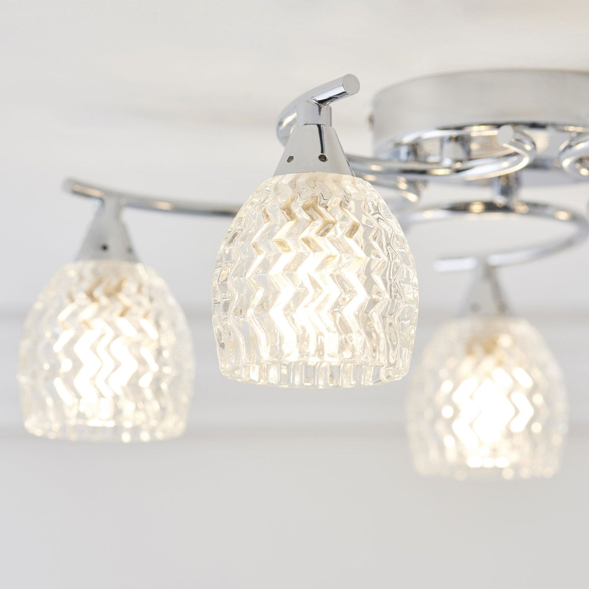 Close-up of three illuminated, clear, textured glass shades and chrome arms of a ceiling light fixture.
