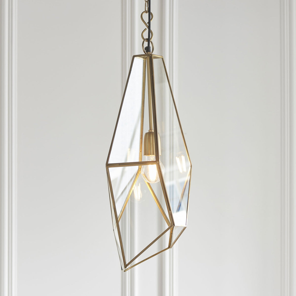 Geometric pendant light with clear glass panels and a brass frame, hanging against a white paneled wall.