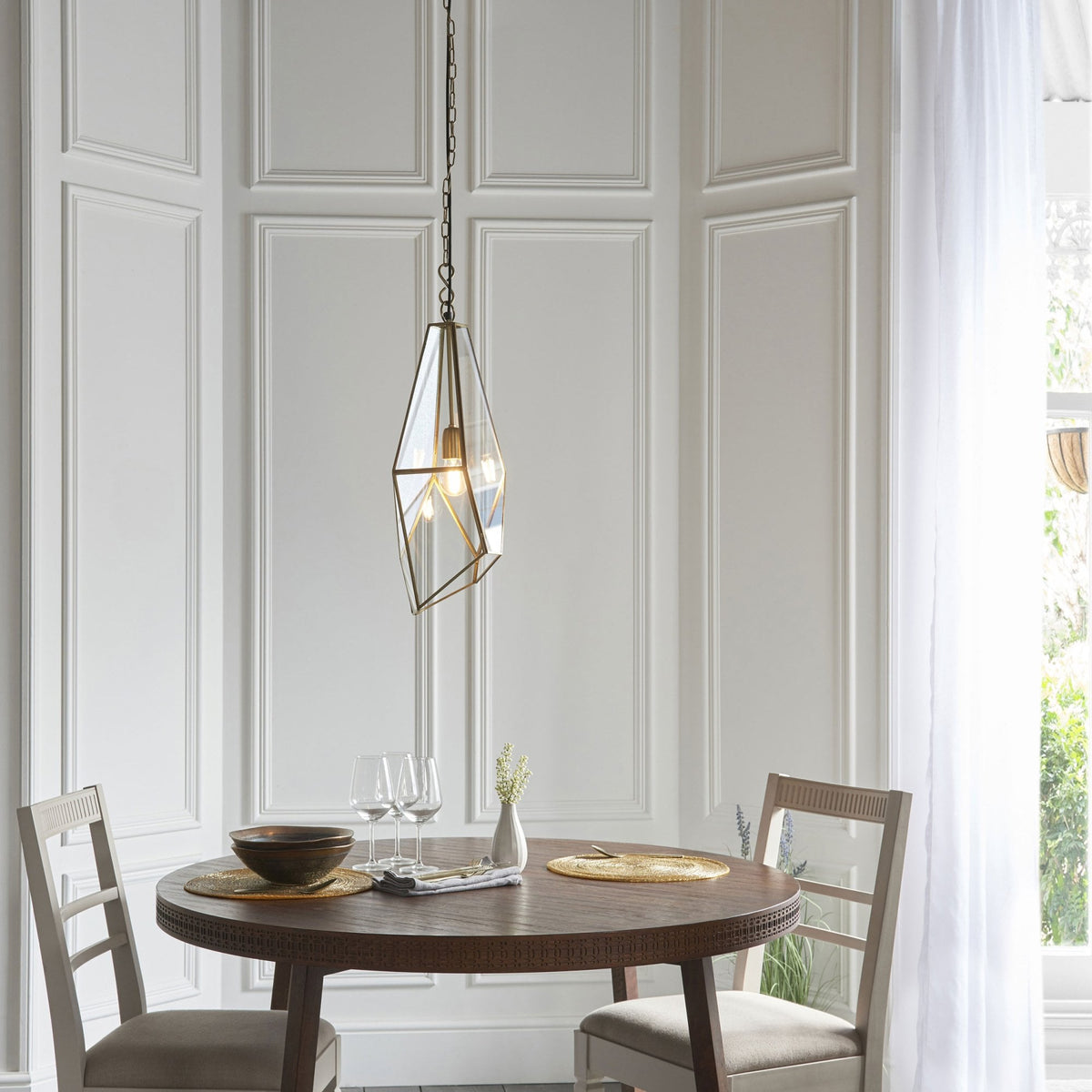 Geometric pendant light hanging above a round dining table with two chairs in a room with white paneled walls.