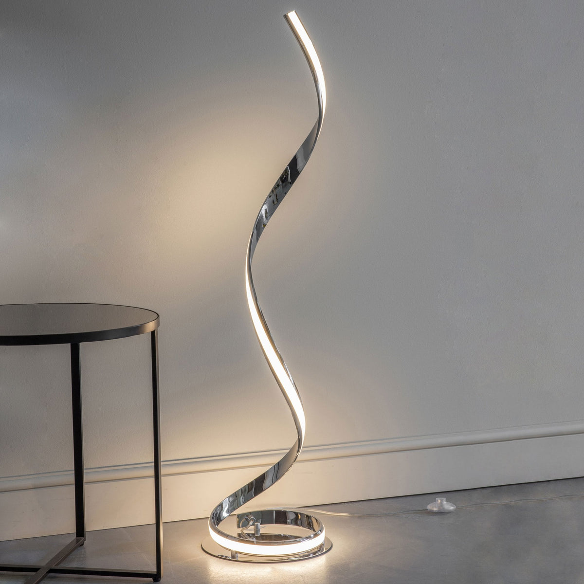 Modern chrome spiral LED floor lamp illuminating a room next to a dark round side table.
