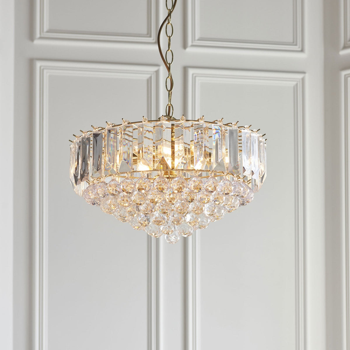 Close-up of a gold and crystal chandelier with a white paneled wall in the background.