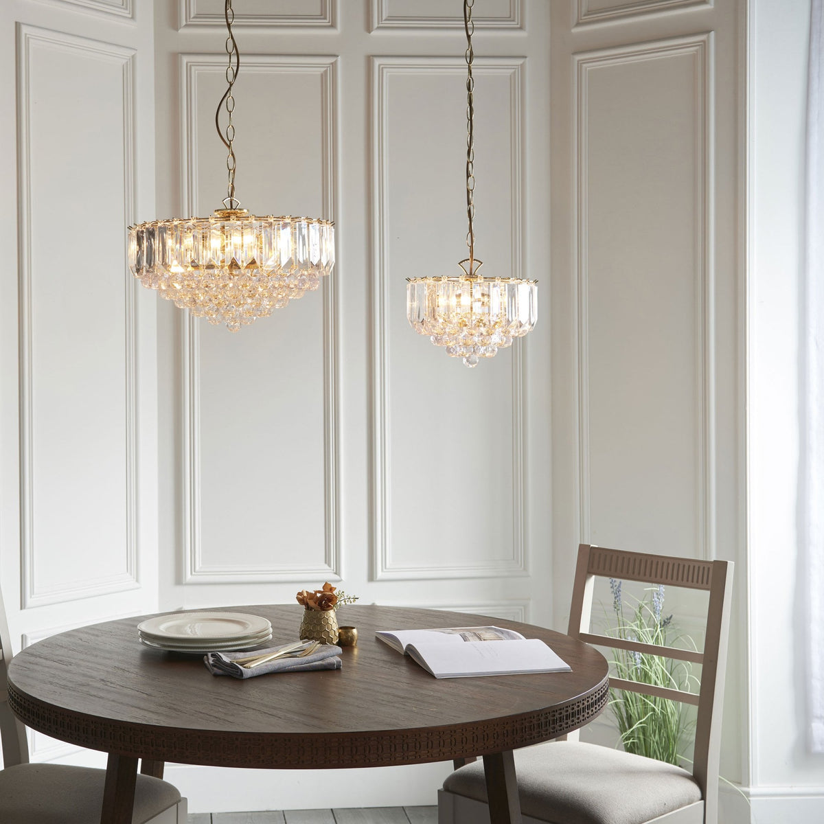 Two gold and crystal chandeliers hanging above a round dining table in a room with white paneled walls.