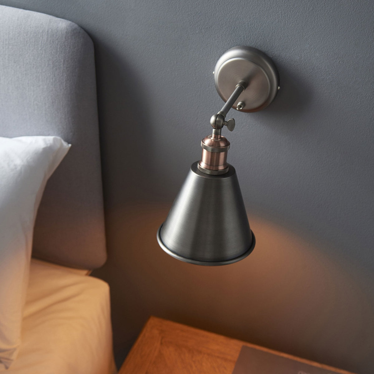Wall light with a dark metal shade and a copper accent, casting a warm glow on a gray wall next to a bed.