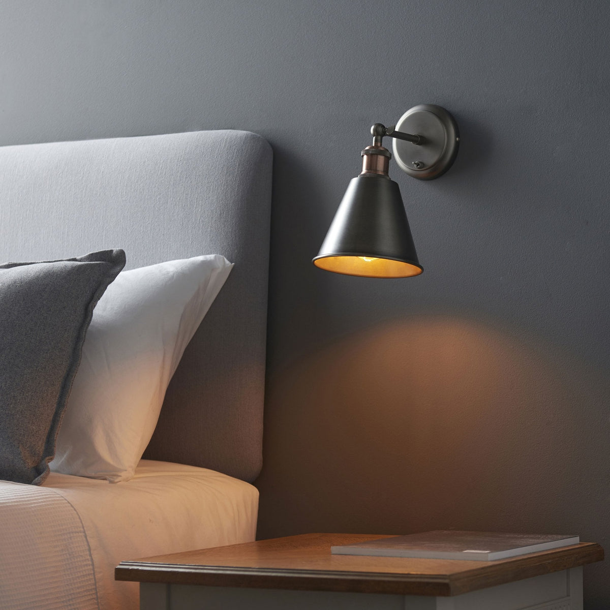 Wall light with a dark metal shade and a copper accent, casting a warm glow on a gray wall next to a bed.