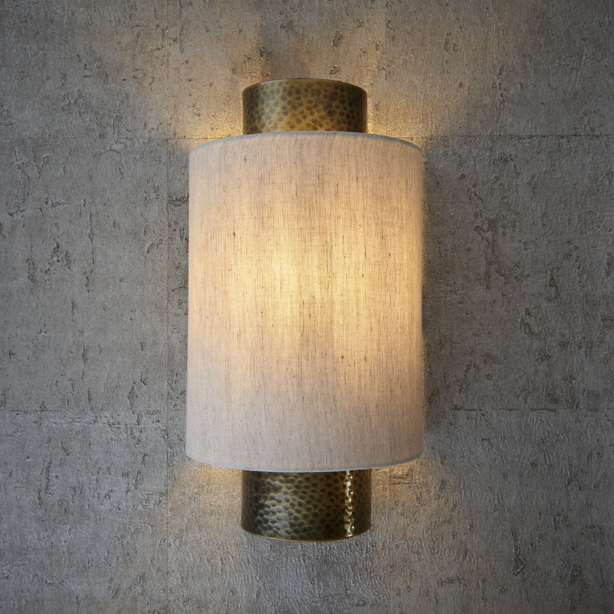 Cylindrical wall light with a textured brass base and top, and a light fabric shade, illuminated on a concrete wall.