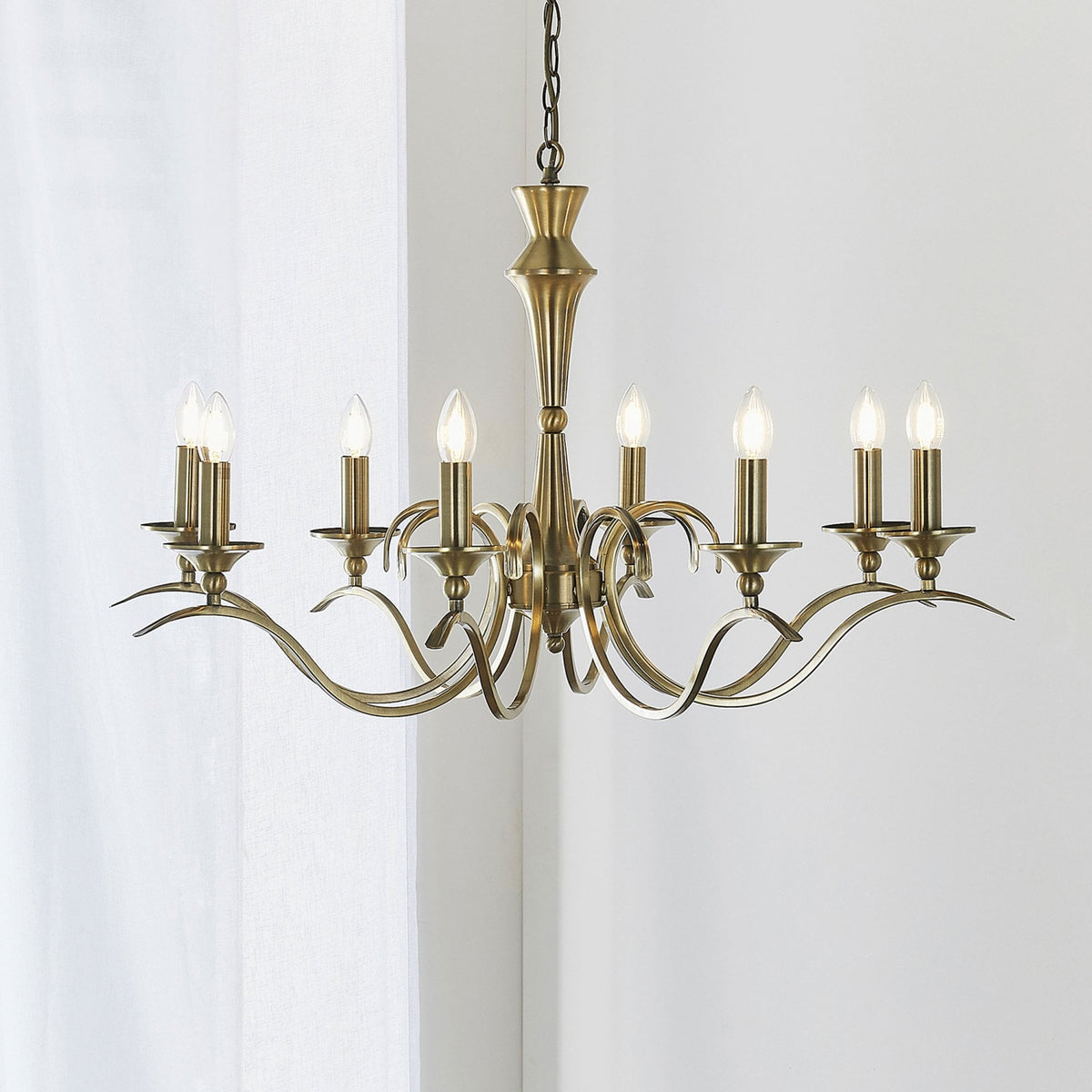 Eight-light antique brass chandelier with curved arms and candle-style bulbs against a white curtain.
