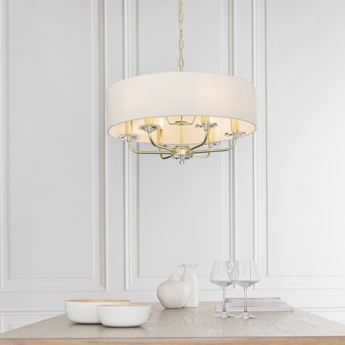 Drum shade chandelier with gold accents and crystal details above a dining table with white decor.