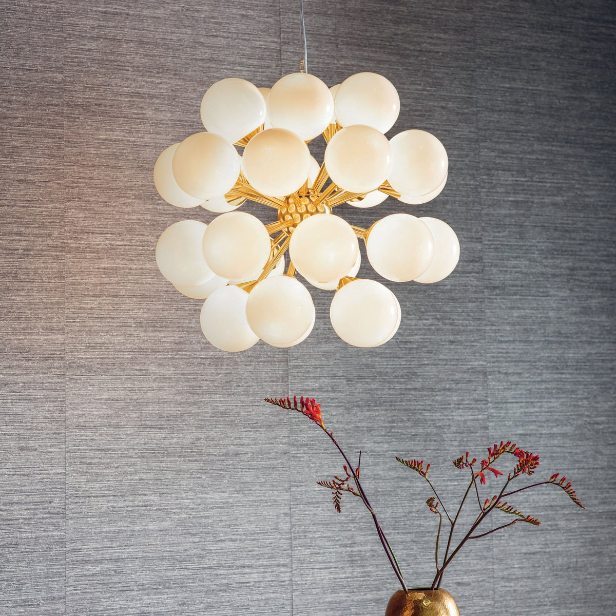 A large, modern chandelier with numerous white spherical lights and a gold frame, hanging above a vase with red flowers.