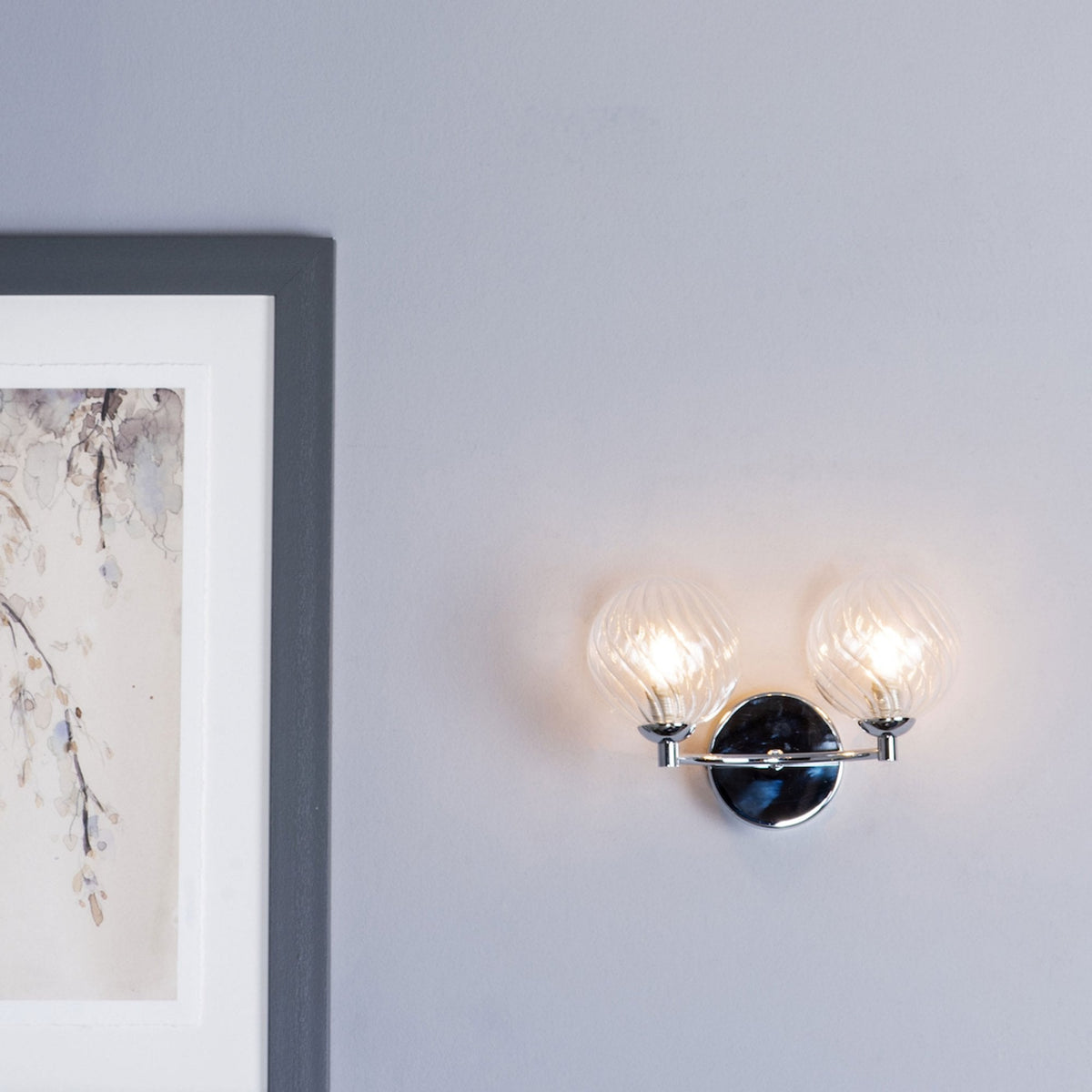 A modern chrome wall light with two clear, swirled glass shades illuminates a light gray wall next to a framed picture.