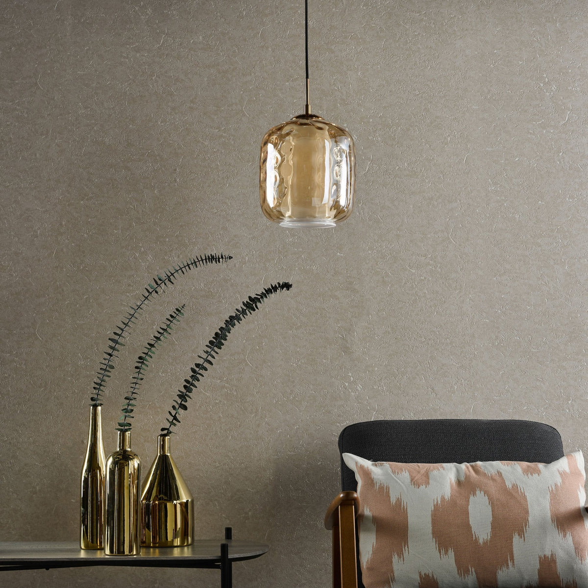 A textured beige wall with a golden pendant light, three golden vases with eucalyptus, and a chair with a patterned pillow.