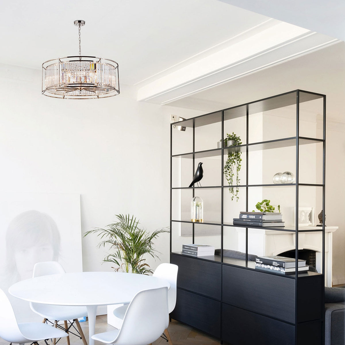 A modern dining room with a crystal chandelier, white table, and black shelving unit.