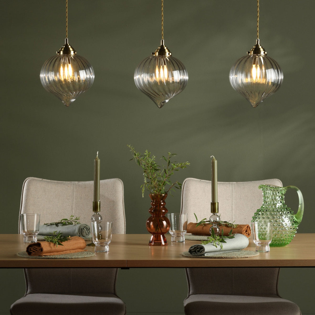 Three glass pendant lights illuminate a dining table set with candles, a vase, and rolled napkins.