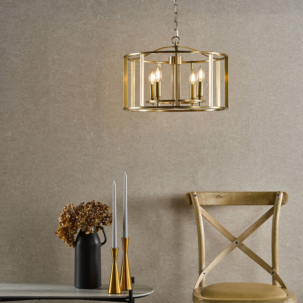 A gold chandelier hangs above a table with a black vase, dried flowers, and two gold candle holders.