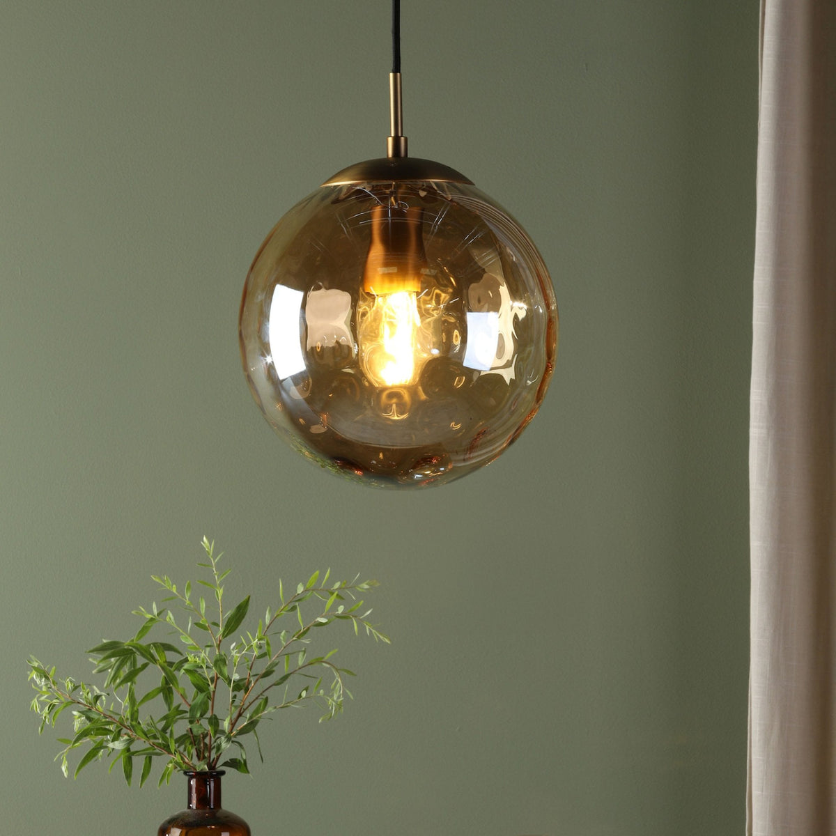 A close-up of a glass pendant light with a warm glow, hanging against a green wall.