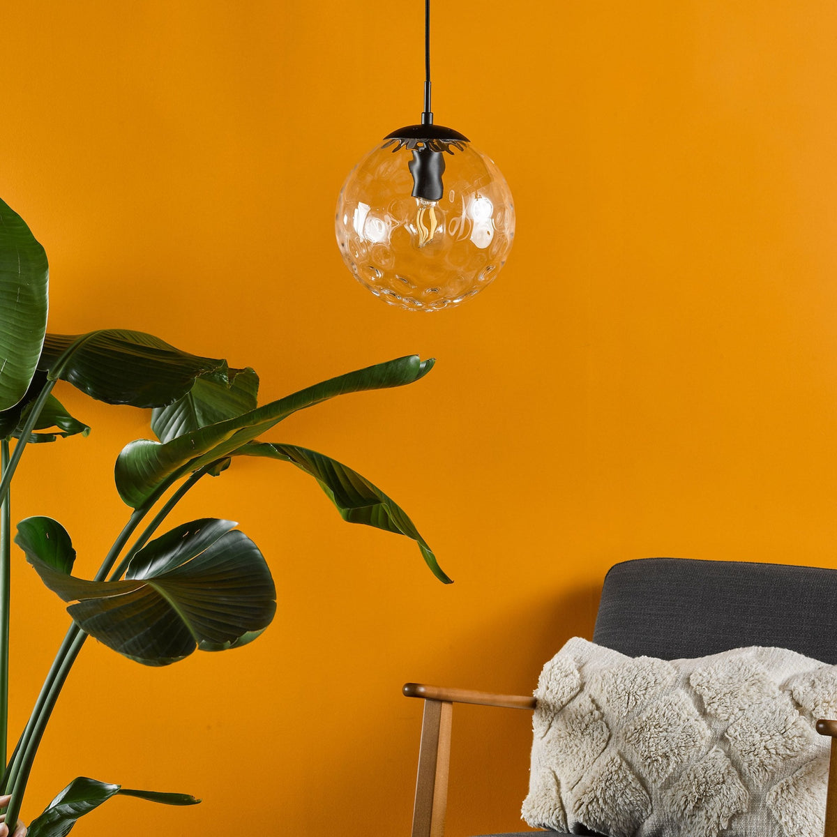 A clear, textured glass globe pendant light hangs above a dark gray armchair with a cream-colored pillow.
