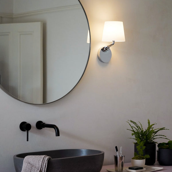 Round mirror above a dark grey basin with black wall-mounted tap, a wall light, and potted plants