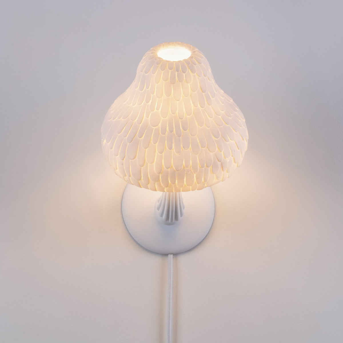 A white mushroom-shaped wall lamp with a textured shade illuminates a white wall.
