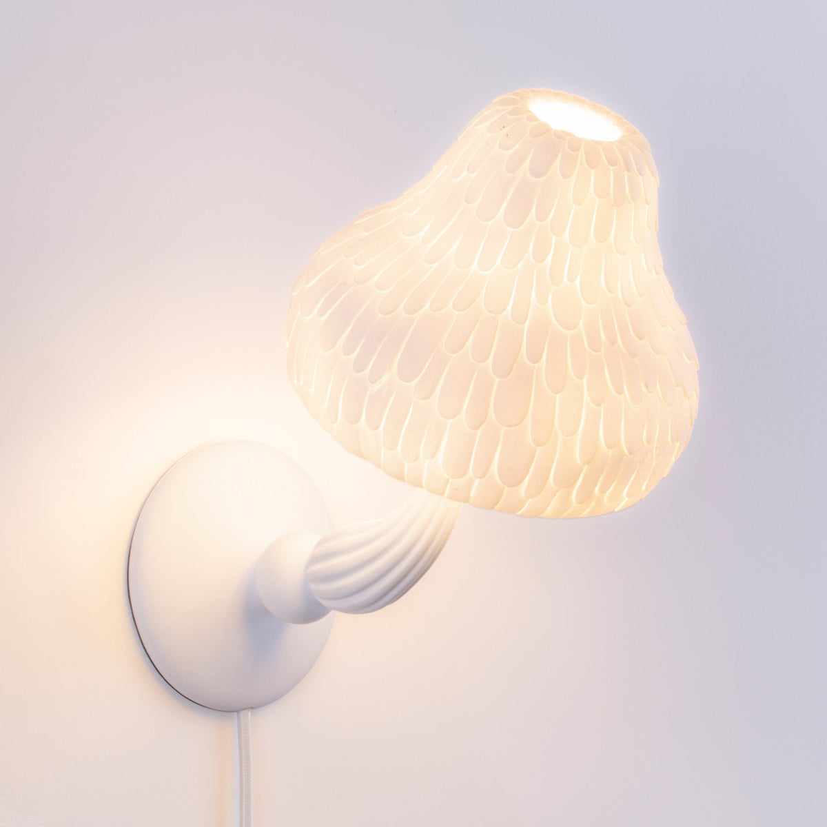 A white mushroom-shaped wall lamp with a textured shade glows brightly on a white wall.