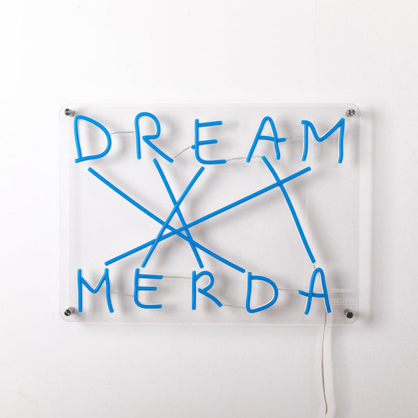 Blue neon sign reads 'DREAM MERDA' with lines connecting letters, on clear backing.