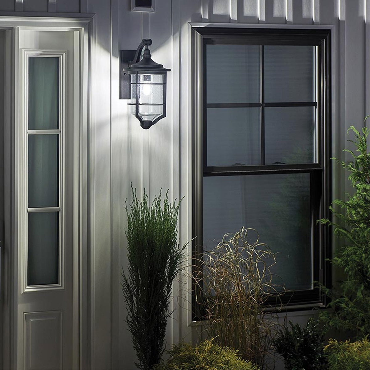 An outdoor wall light with a classic design illuminates a white house at dusk.