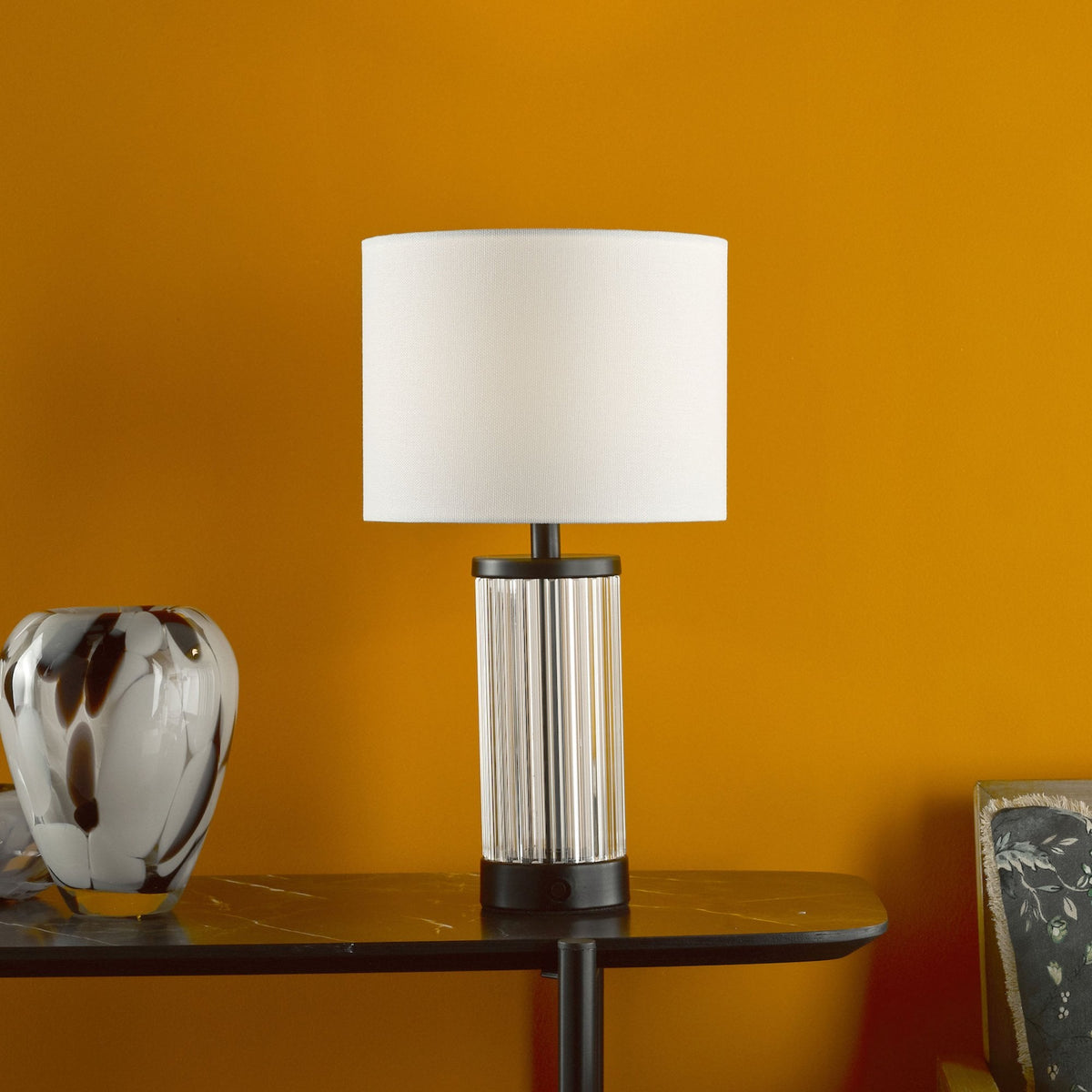 A white lampshade on a fluted glass lamp base sits on a black marble table against an orange wall.