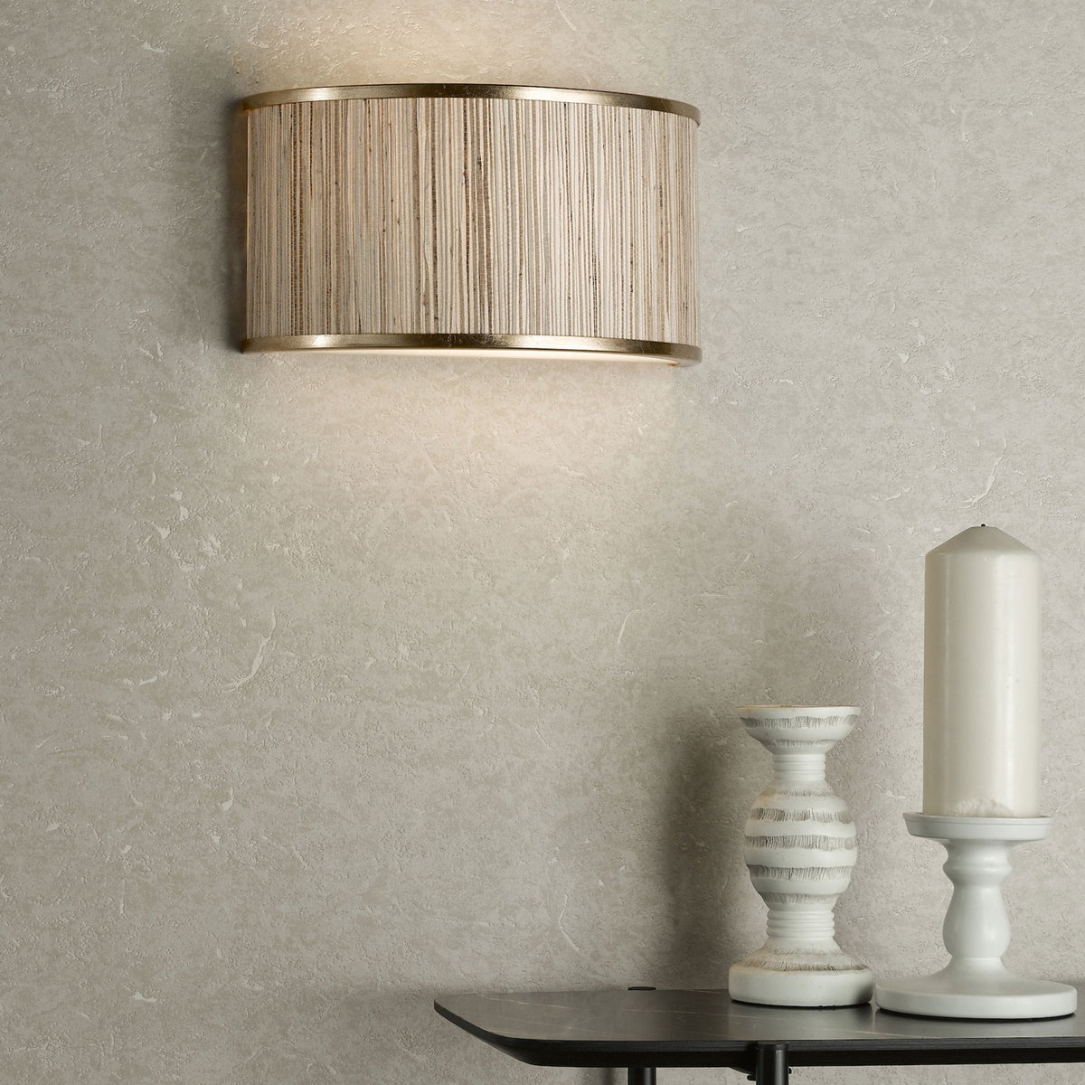 Textured wall with a gold and woven sconce, a white candle, and a decorative white holder on a dark table.