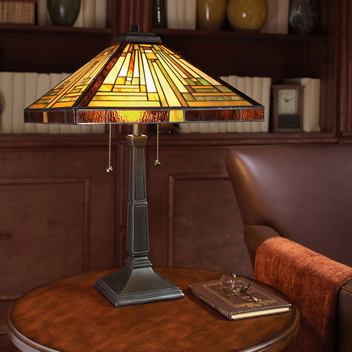 A vintage table lamp with a rectangular stained-glass shade, casting a warm glow in a study.