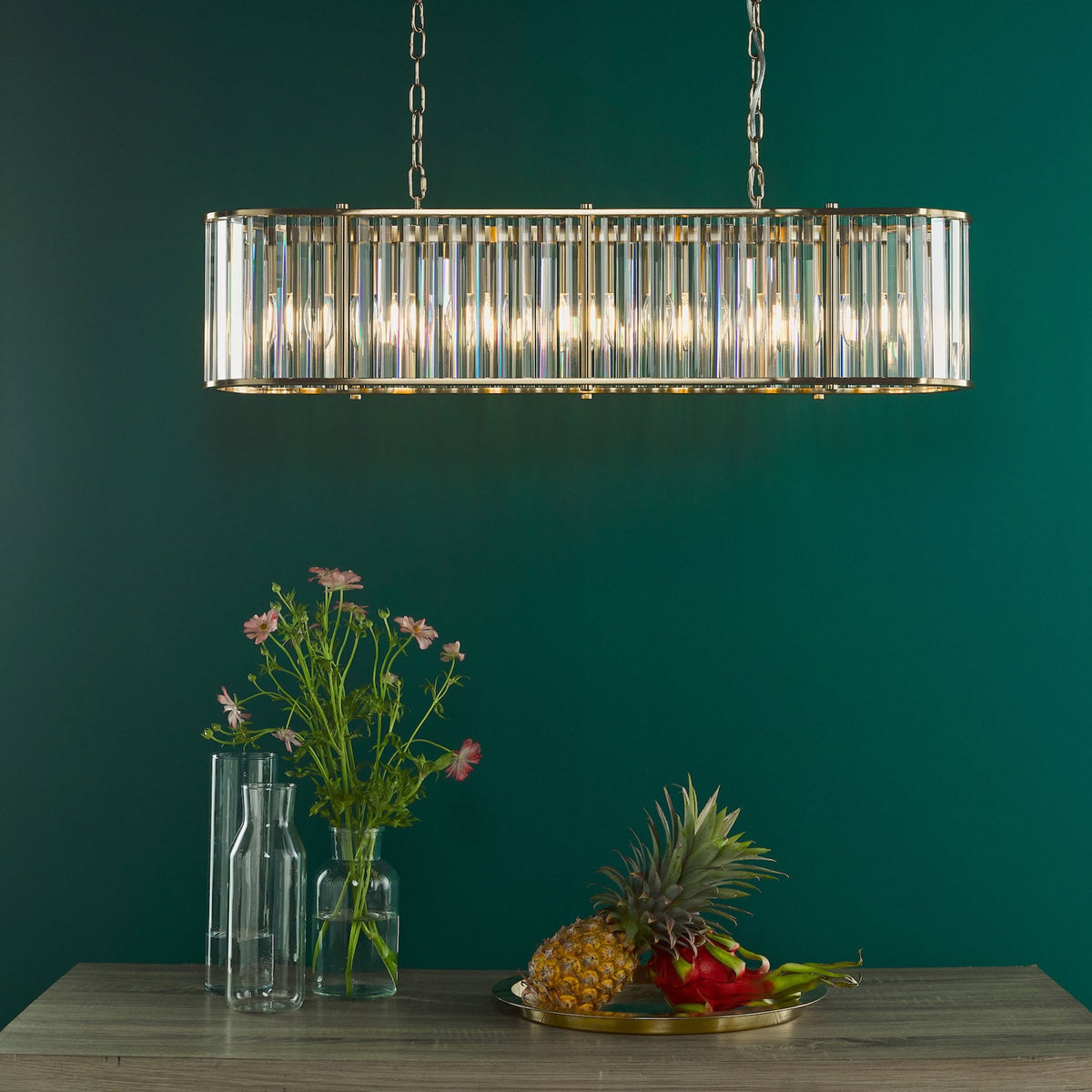 Rectangular crystal chandelier with gold accents, illuminating a dark green wall above a wooden table with flowers and fruit.