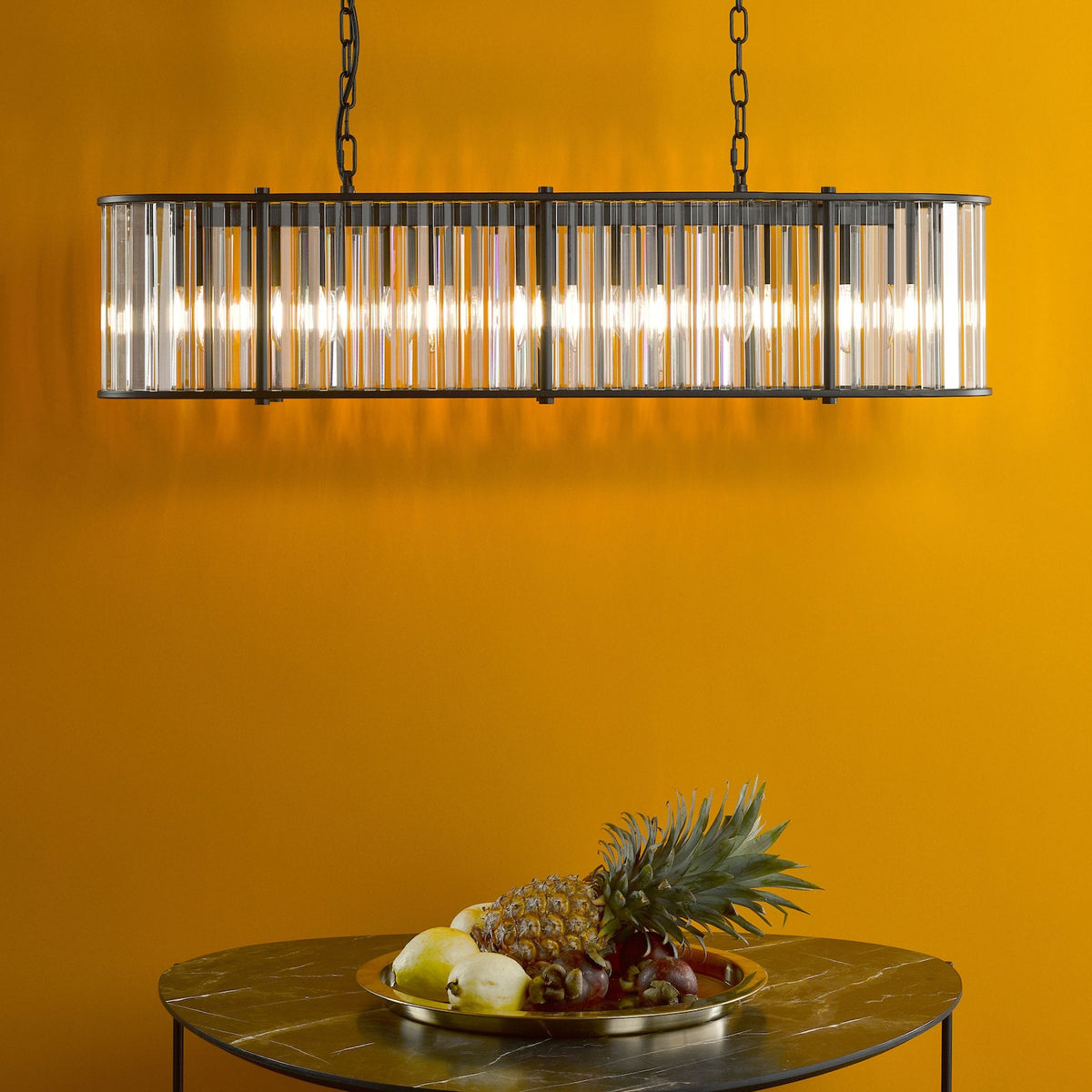 Rectangular chandelier with clear glass rods and a black frame, hanging above a fruit bowl on a marble table.