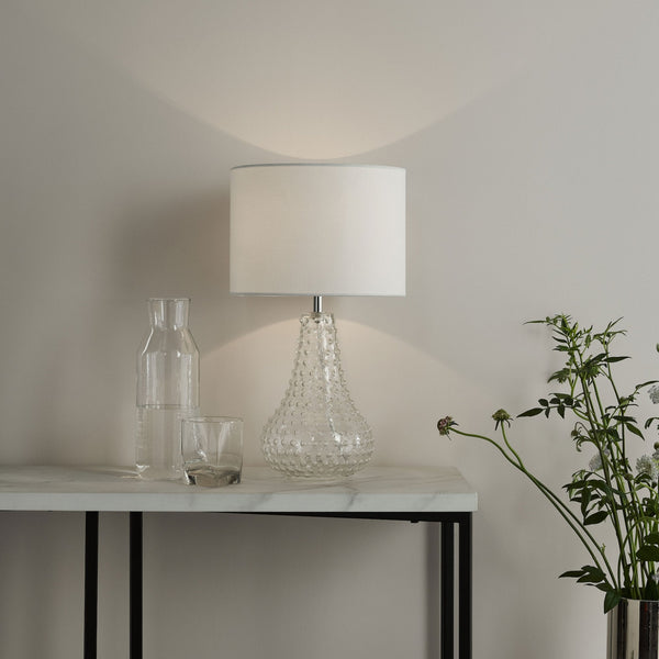 A clear glass lamp with a white shade sits on a marble table next to a glass carafe and a vase of greenery.