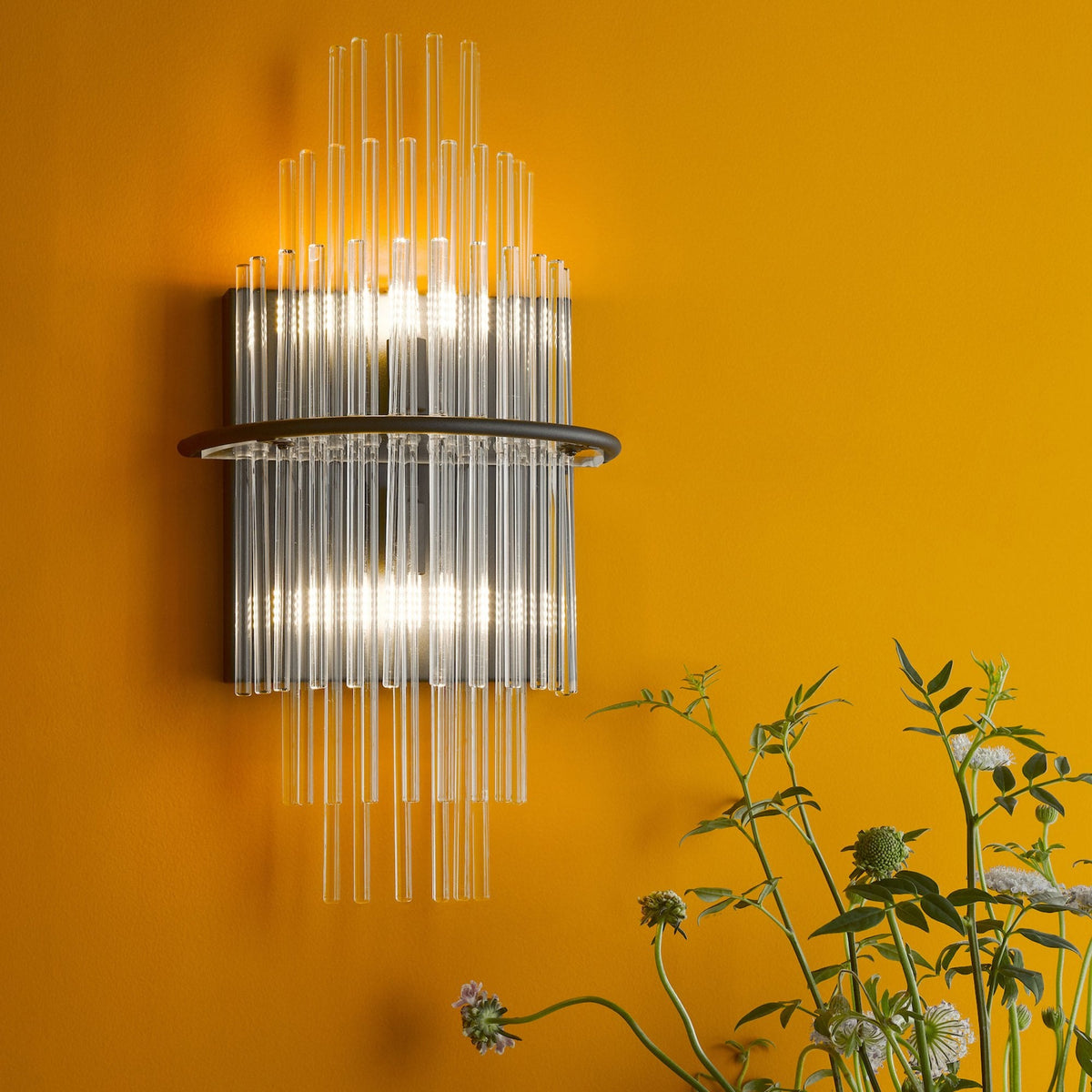 A modern wall sconce with clear glass tubes and a dark metal band, casting light on a vibrant orange wall.