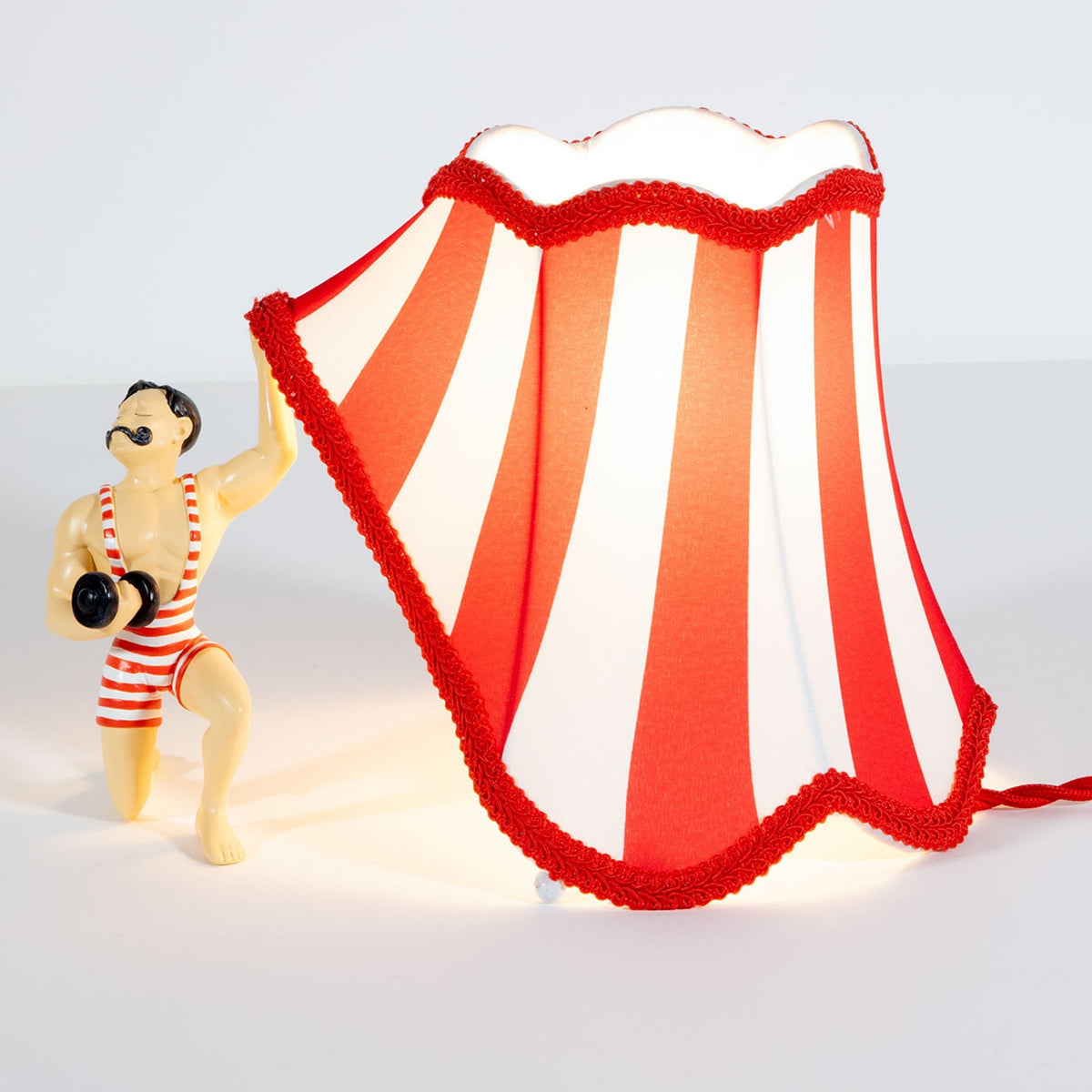 A circus strongman holding up a red and white striped lampshade, creating an ambient glow.