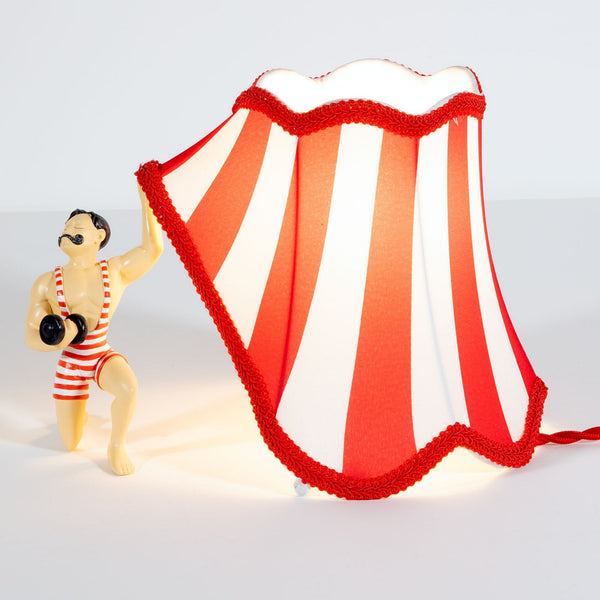 A circus strongman holding up a red and white striped lampshade, creating an ambient glow.
