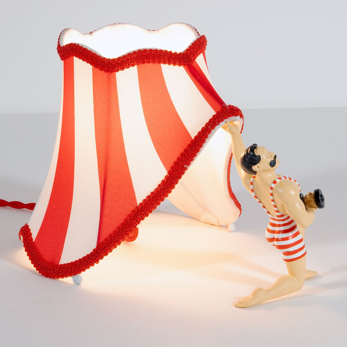 A circus strongman from the left, holding up a red and white striped lampshade.