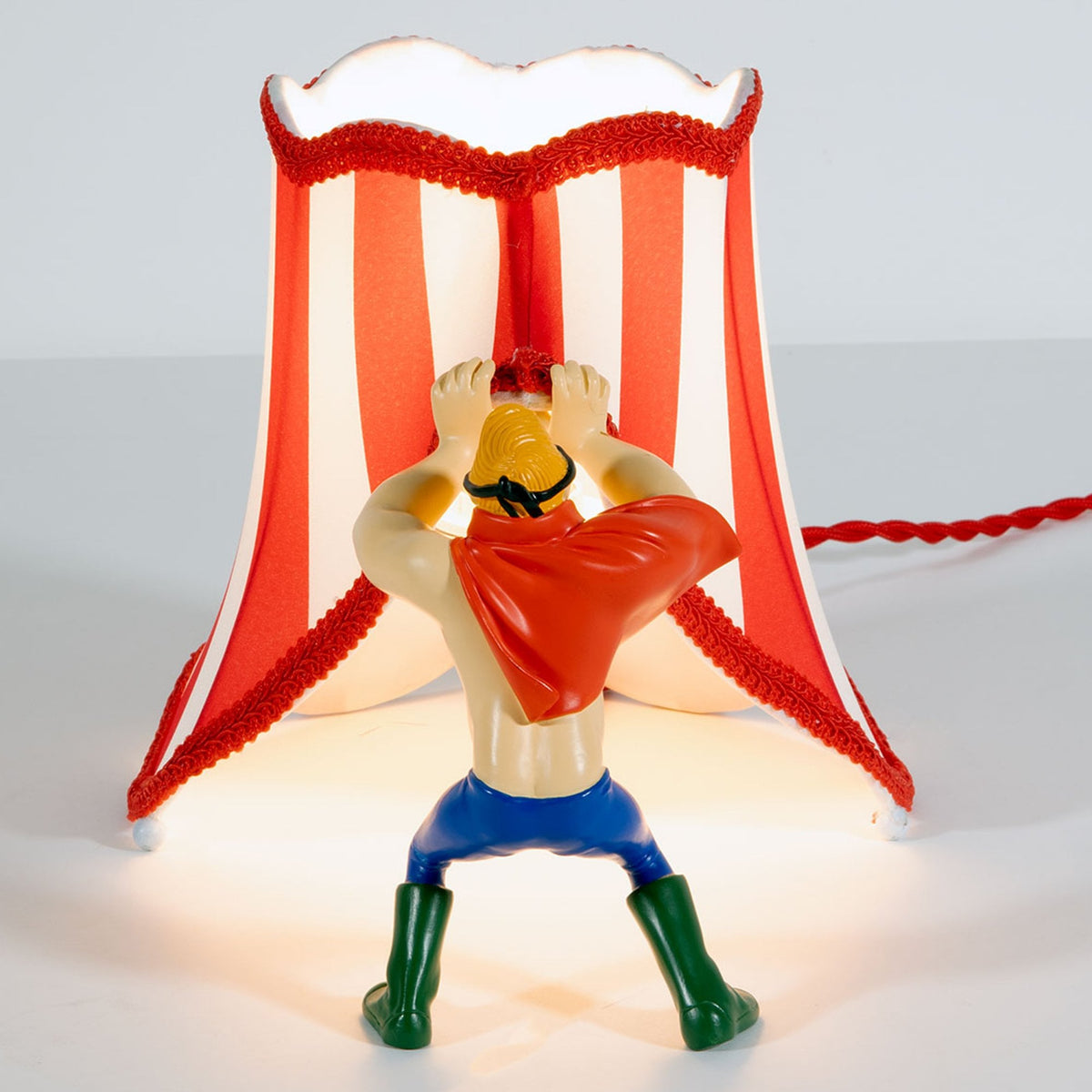 A figurine holds up a red and white striped lampshade, standing beneath it like a tent.