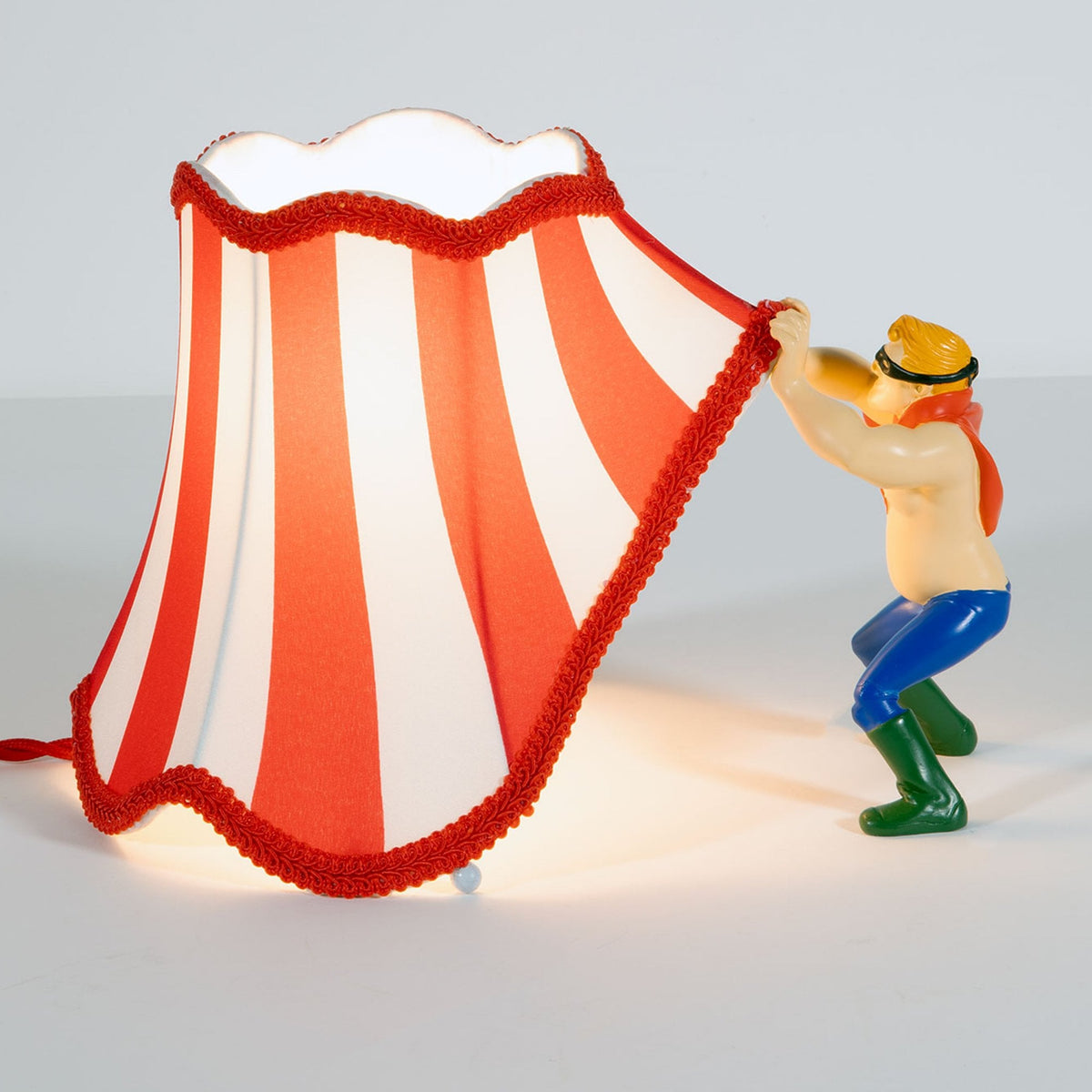 A figurine holds up a red and white striped lampshade, resembling a circus tent, side view.